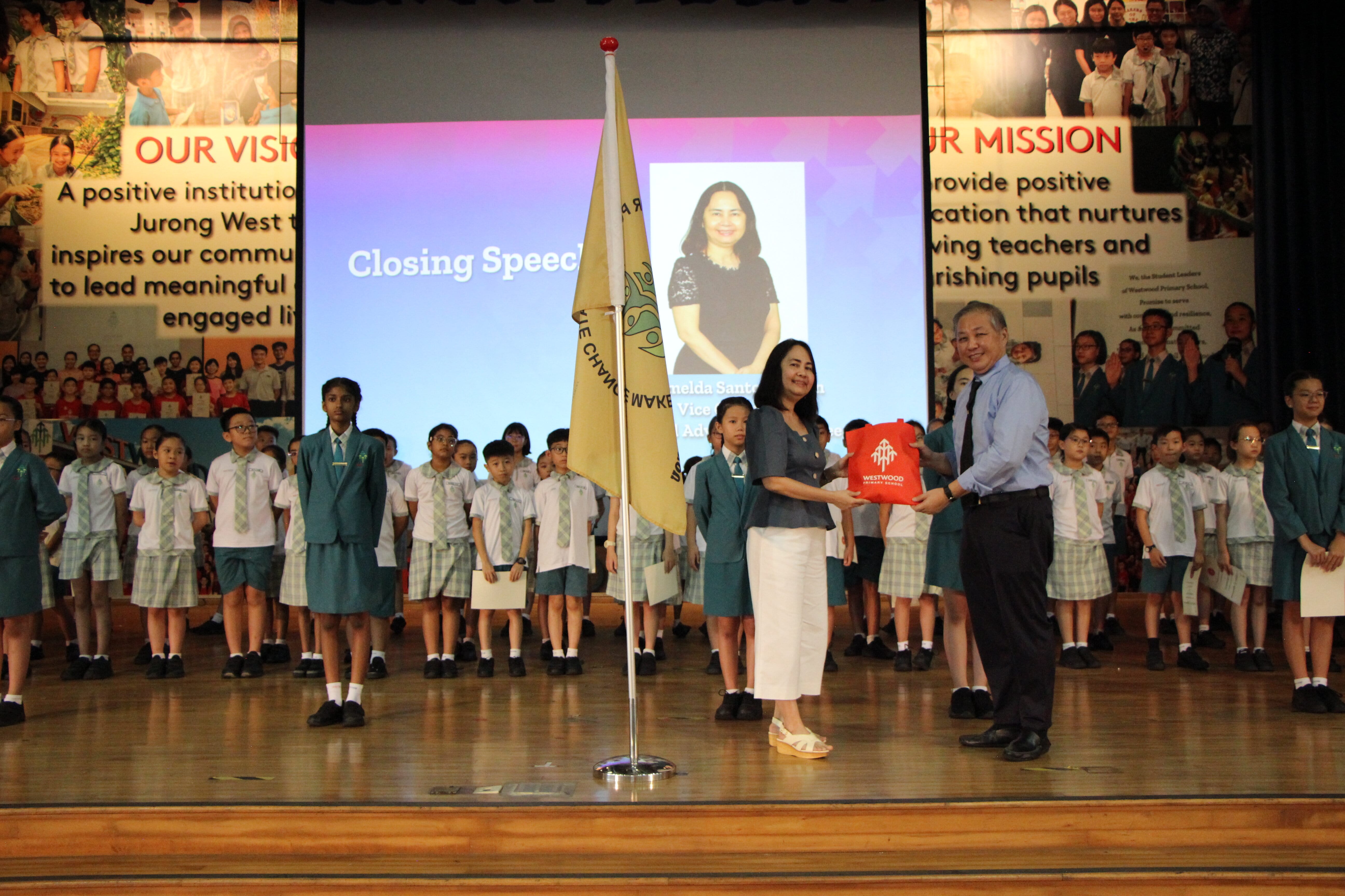 Two adults exchange a red gift bag on stage at a school closing speech ceremony, with uniformed students standing behind them.